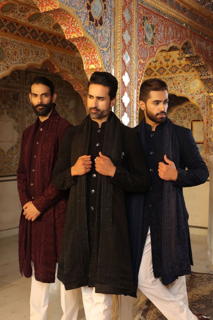 Three men in traditional attire pose in an ornately decorated room, exuding elegance.